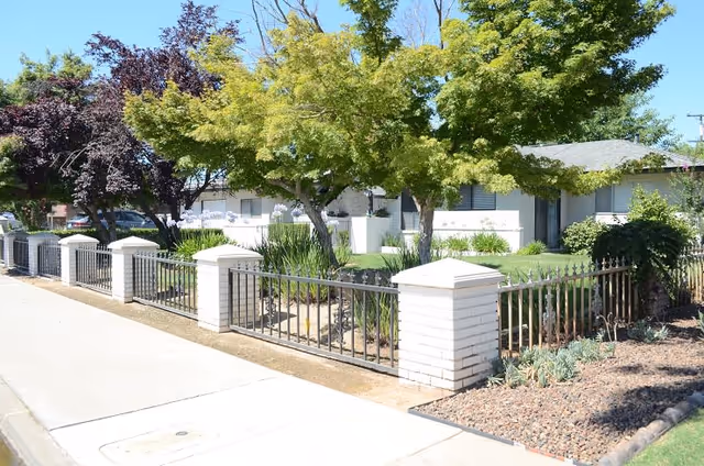 Exterior view of a single-story residential building with white brick pillars and black metal fencing surrounding a landscaped yard with green grass, trees, and shrubs under a clear blue sky.