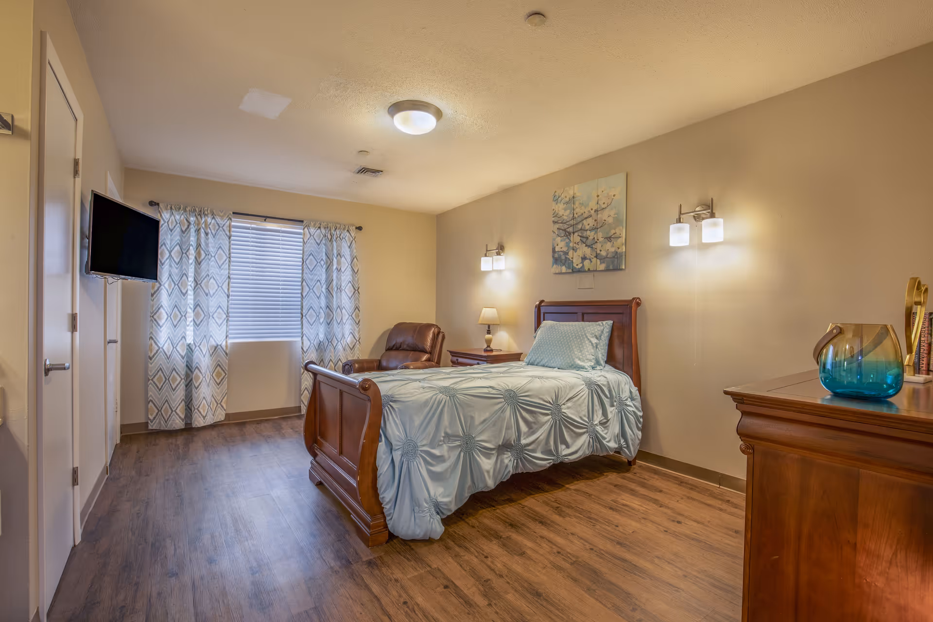 A senior living bedroom with a single wooden bed covered with a light blue comforter and pillow. The room has wooden flooring, a brown leather armchair, a wooden nightstand with a lamp, and a wooden dresser with a blue glass vase. There is a window with patterned curtains and a wall-mounted TV on the left side. Two wall-mounted light fixtures and a ceiling light illuminate the room. A painting with floral artwork hangs above the bed.