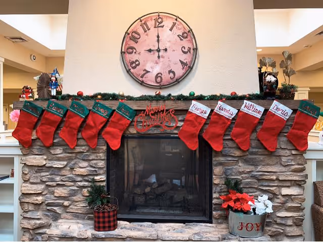 A stone fireplace decorated for Christmas with red stockings hanging from the mantel. Above the mantel is a large round clock showing the time as 11:55. The mantel is adorned with green garland and a red 'Merry Christmas' sign. There are small plants on either side of the fireplace, including a red and white poinsettia in a metal container labeled 'JOY'.