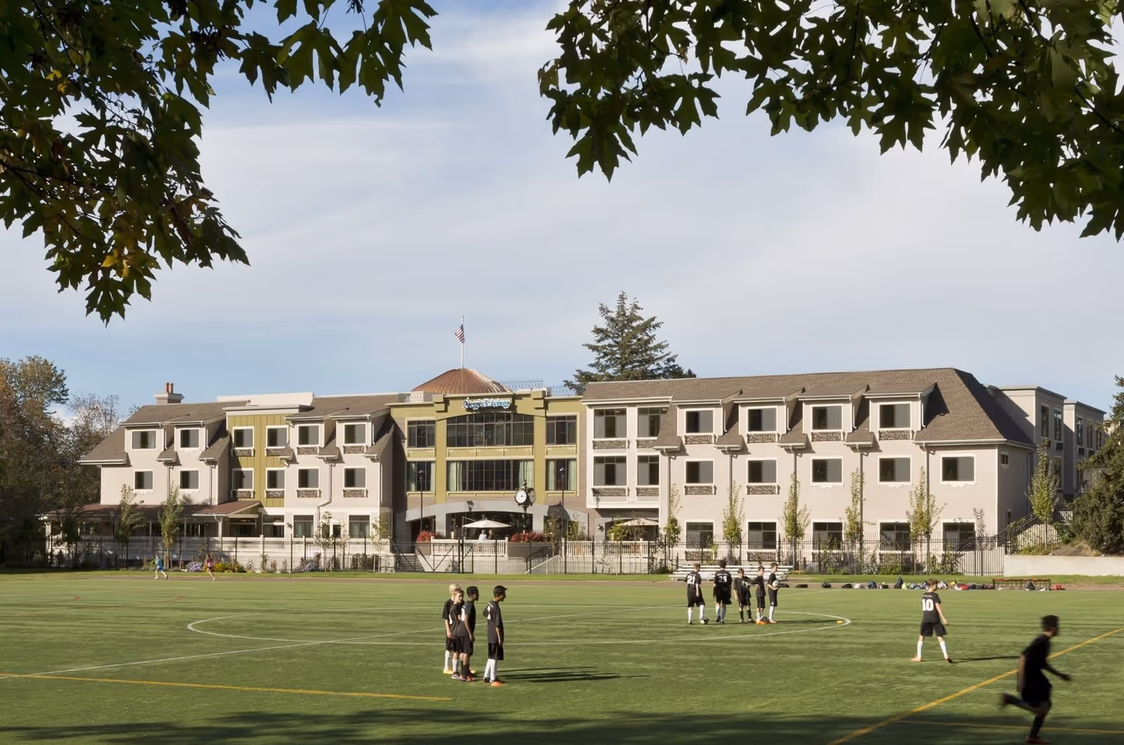 View of a large multi-story building with a green lawn in front where children are playing soccer. The building has multiple windows and a central section with a clock and an American flag on top. Trees frame the image from above.