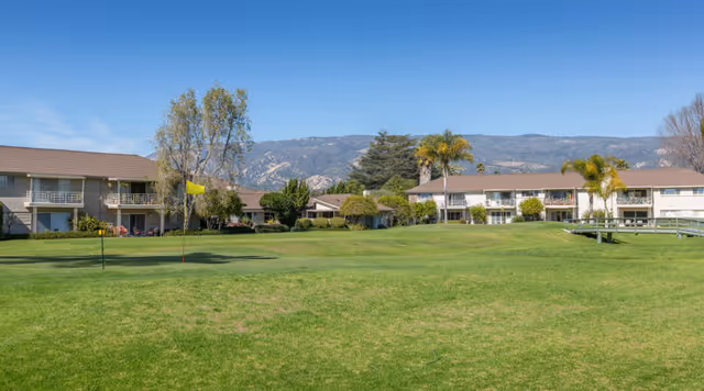 Grassy lawn with a small putting green and flag in front of two-story residential buildings, palm trees, and mountains in the background.