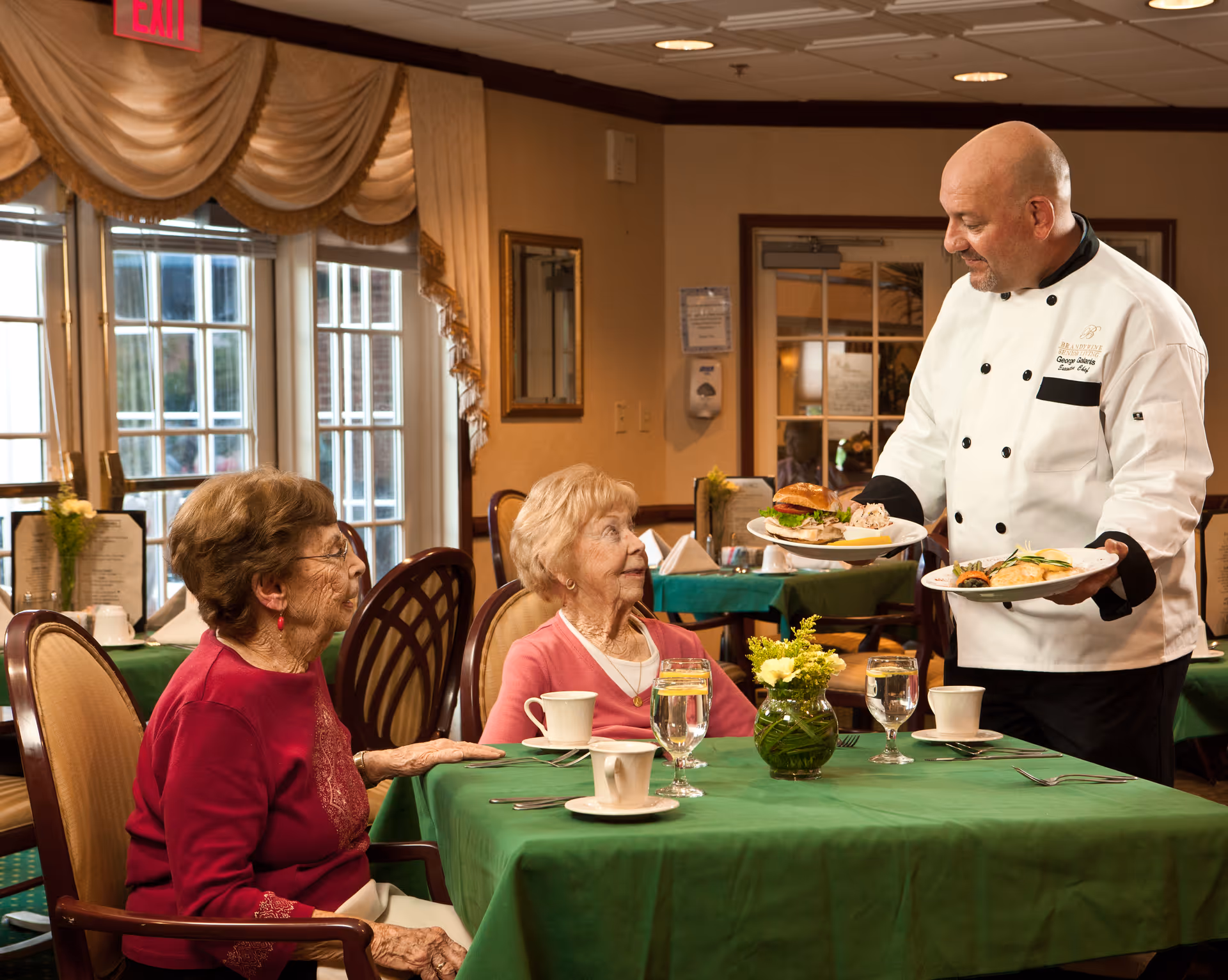 Two elderly women seated at a dining table with green tablecloths in a well-lit dining room. A chef in a white uniform is serving them plates of food. The table is set with cups, glasses of water, and a small vase with yellow flowers. Large windows with draped curtains are visible in the background.