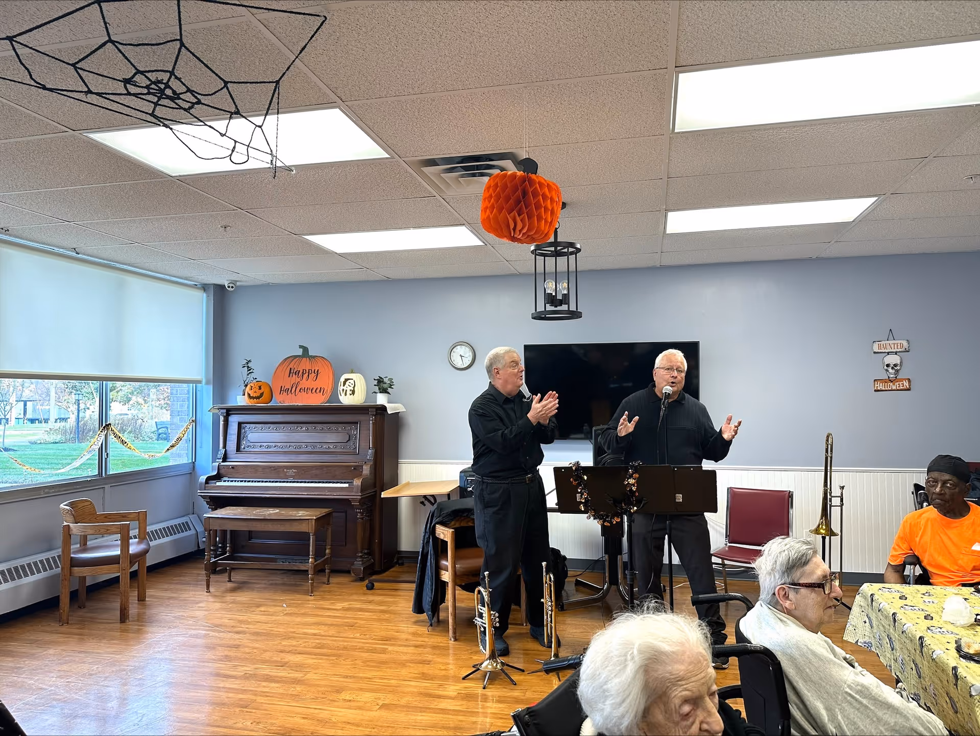 A group of elderly people seated around a table in a common room decorated for Halloween. Two men are standing near a piano and a microphone, one speaking and the other clapping. The room has wooden floors, large windows, and Halloween decorations including pumpkins and a hanging orange paper lantern.