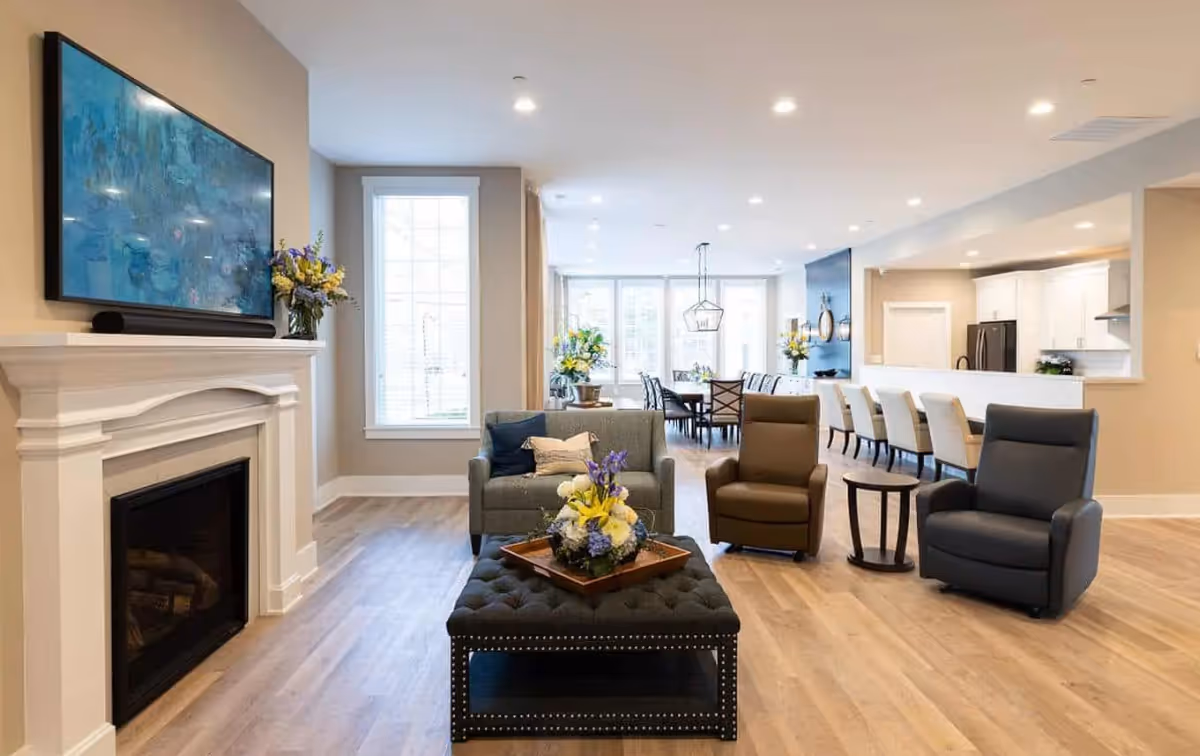 A bright and spacious living area in a senior living facility featuring a white fireplace with a mounted TV above it, a gray loveseat with pillows, two armchairs, a black tufted ottoman with a flower arrangement, and a dining area with a long table and chairs in the background. The room has large windows, light wood flooring, and recessed ceiling lights.