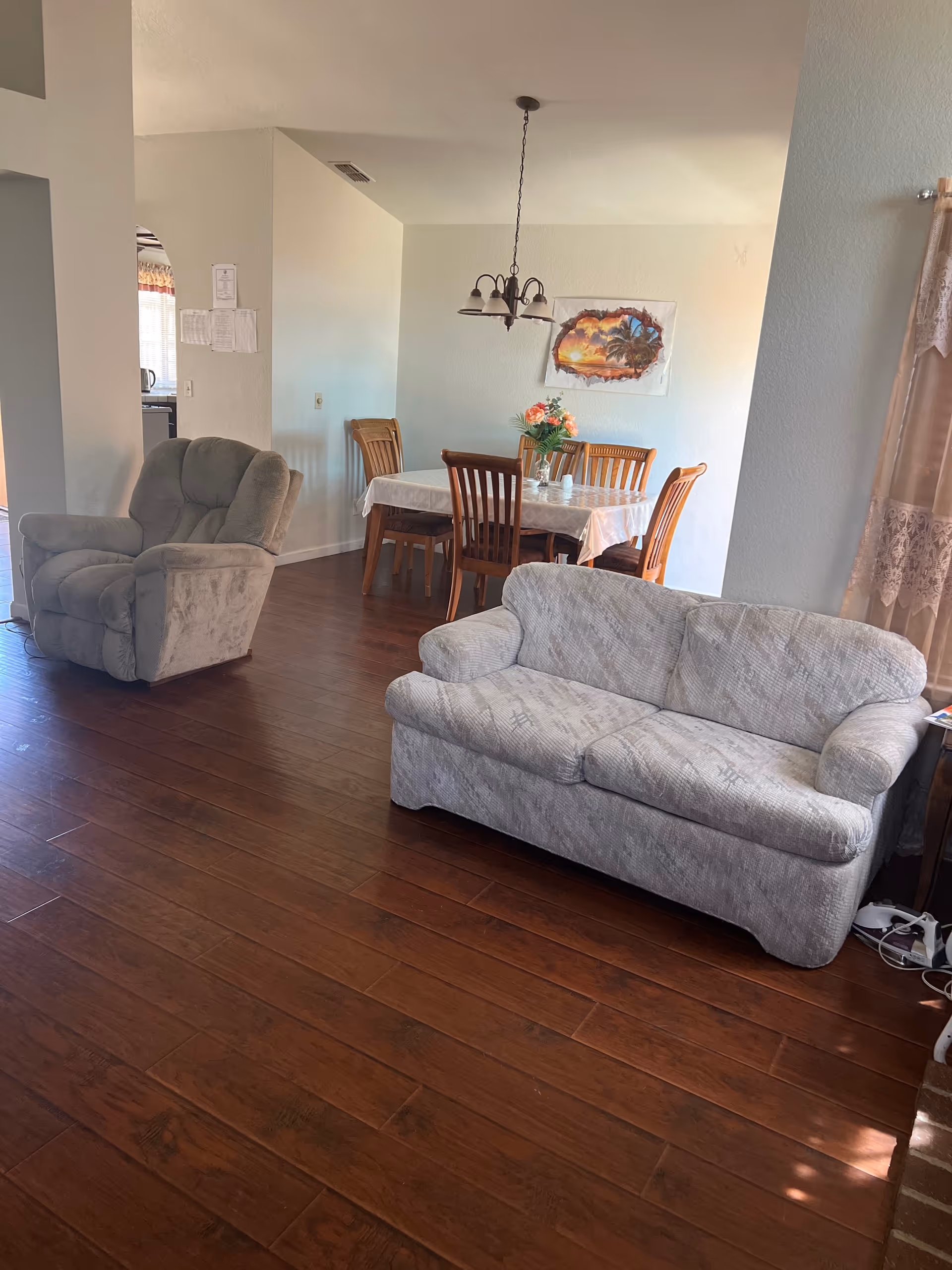 A living room and dining area with wooden flooring. The living room has a light gray sofa and a matching recliner chair. The dining area features a wooden table with six chairs and a vase with flowers on the table. A chandelier hangs above the dining table, and there is a painting on the wall. Light filters in through a window with lace curtains on the right side.