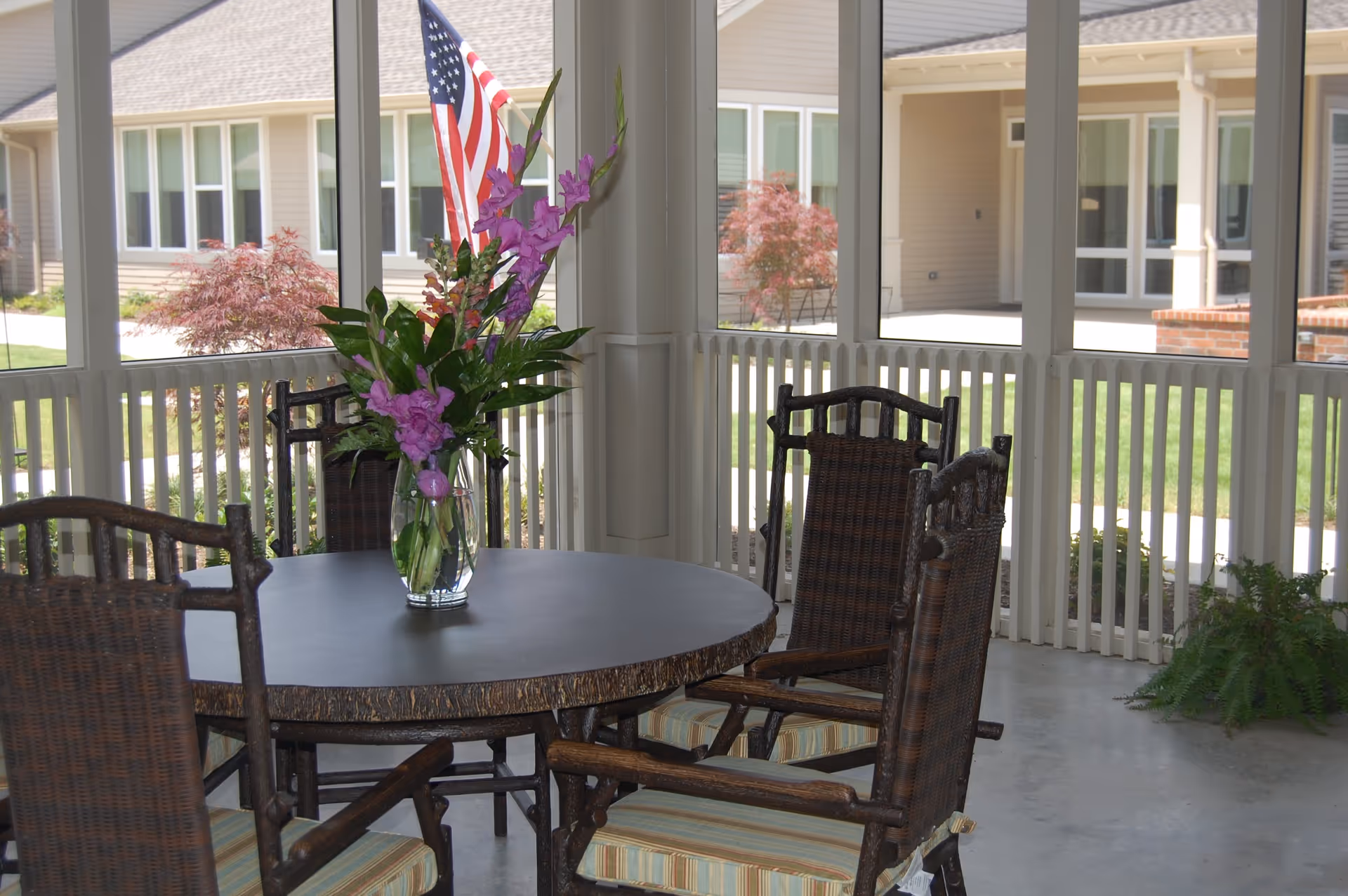 A screened-in porch area with a round table and four wooden chairs with striped cushions. A vase with purple flowers and an American flag is placed on the table. Outside the porch, there is a view of a building with beige siding, multiple windows, and some landscaping including small trees and grass.