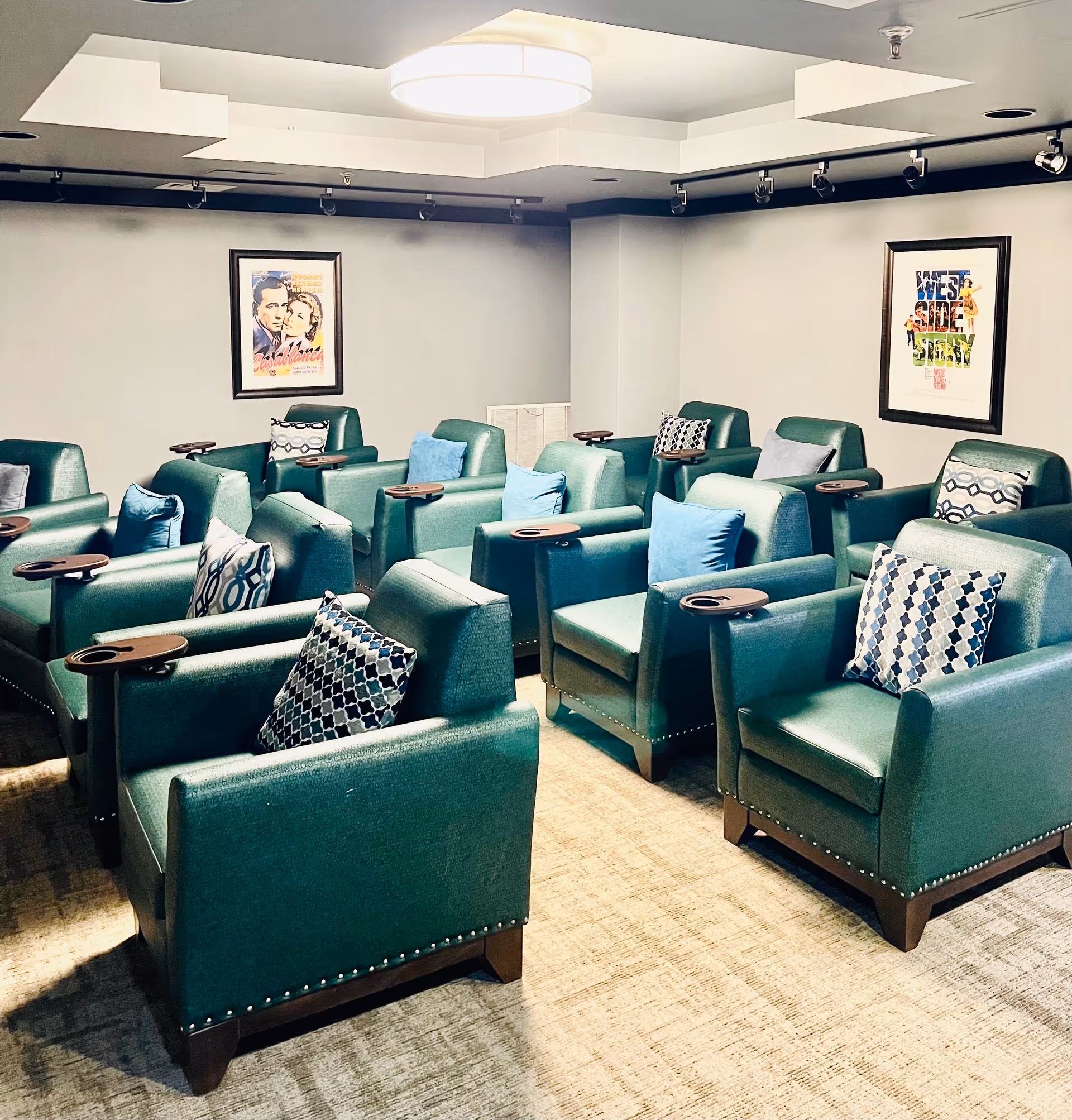 A small theater room with three rows of green armchairs, each with a small attached wooden tray and decorative pillows. The walls are light gray with two framed movie posters, one for Casablanca and one for West Side Story. A large ceiling light fixture illuminates the room.