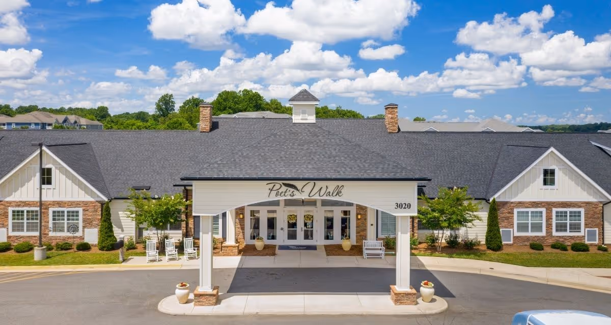 Front exterior view of Poet's Walk Warrenton, A Memory Care Community, showing a single-story building with a covered entrance, white pillars, and a sign with the facility name. The building has a dark shingled roof, brick and siding walls, and landscaping with small trees and bushes under a partly cloudy blue sky.