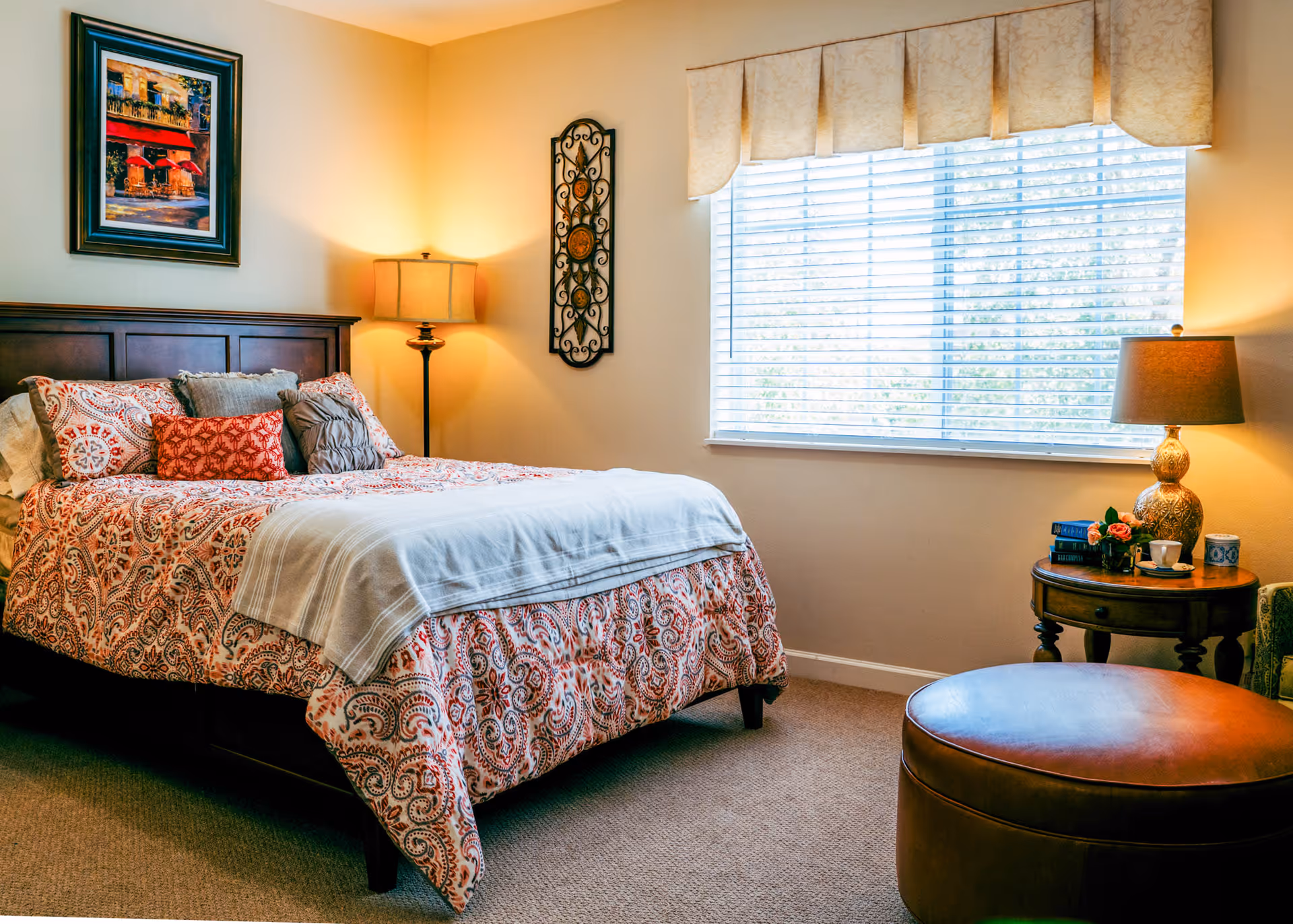 A cozy bedroom with a large bed featuring a patterned orange and white bedspread and multiple pillows. There is a wooden headboard, a floor lamp beside the bed, a framed painting on the wall, and a decorative wall piece. A window with blinds and a valance lets in natural light. A round wooden side table with a lamp, books, a cup, and flowers is next to a brown leather ottoman.