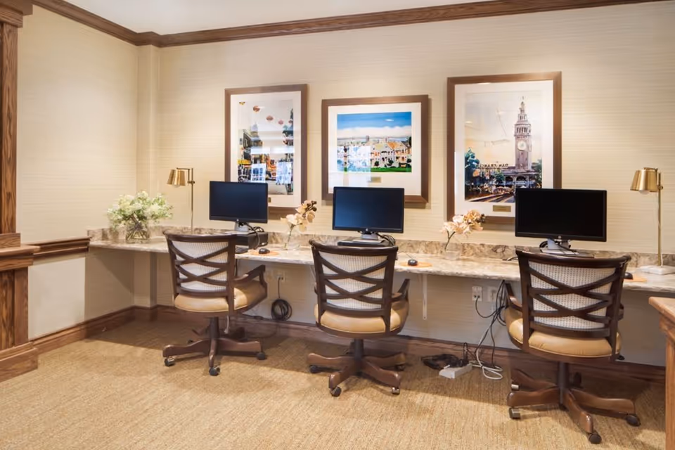 A computer workstation area with three chairs and three desktop monitors on a long marble countertop. The room has beige walls with wooden trim, three framed pictures hanging above the countertop, and decorative flowers placed between the monitors. There are also two brass desk lamps on either end of the countertop.