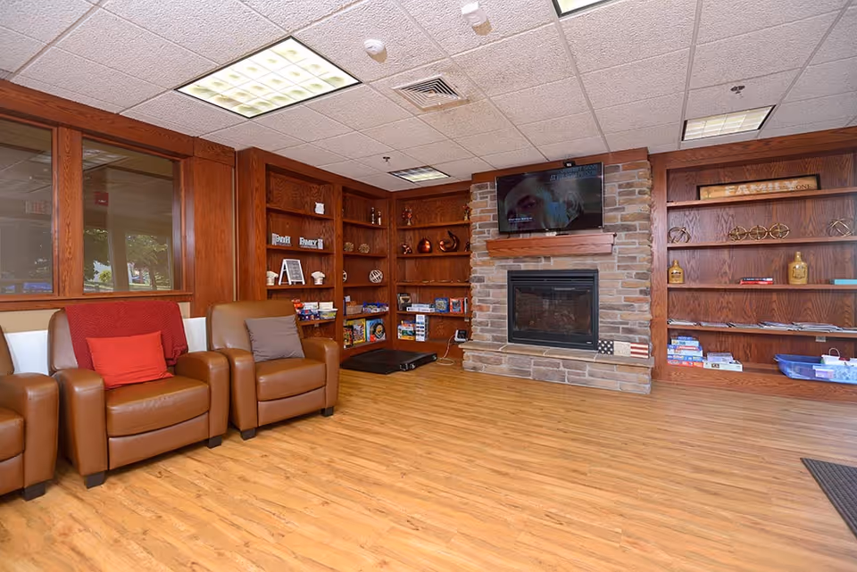 A cozy common area with wooden flooring, brown leather armchairs with red and gray cushions, built-in wooden bookshelves filled with books and decorative items, a stone fireplace with a mounted flat-screen TV above it, and large windows on the left side.