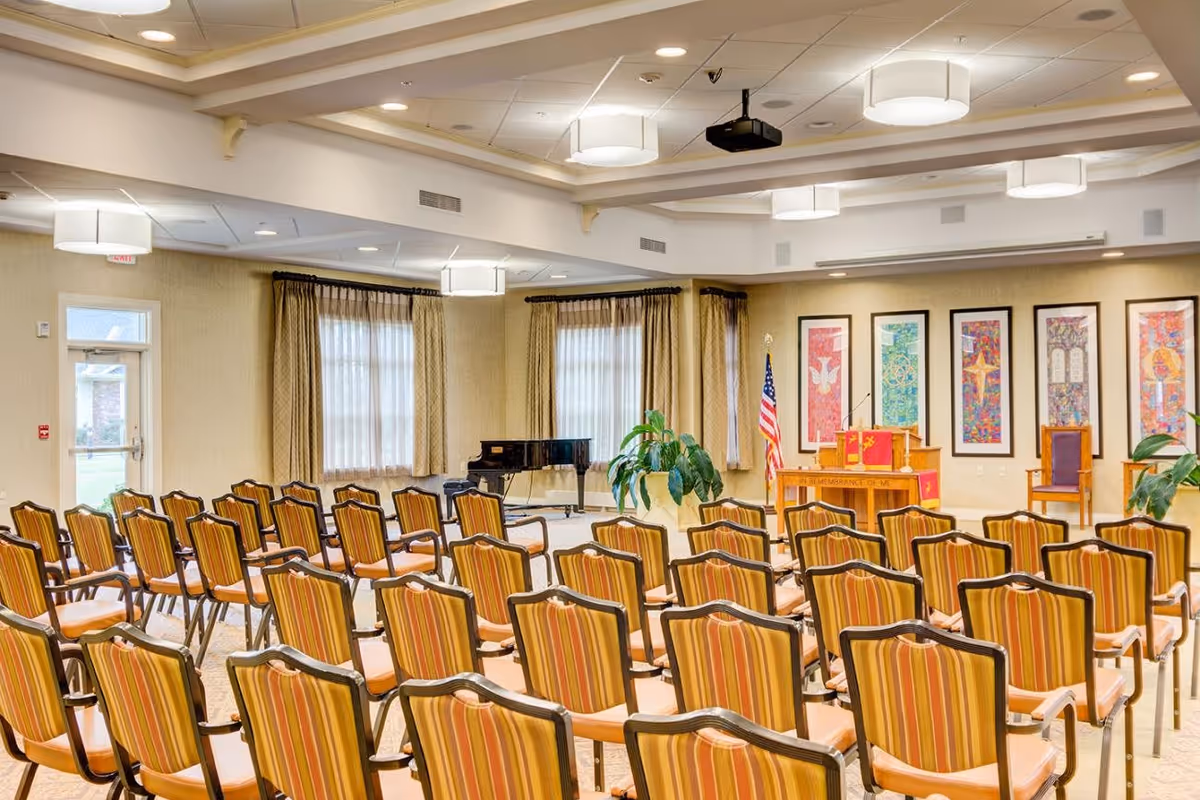 A well-lit chapel or meeting room with rows of striped cushioned chairs facing a wooden podium adorned with religious symbols and an American flag. The room has large windows with curtains, framed colorful artwork on the walls, a grand piano in the corner, and ceiling lights.