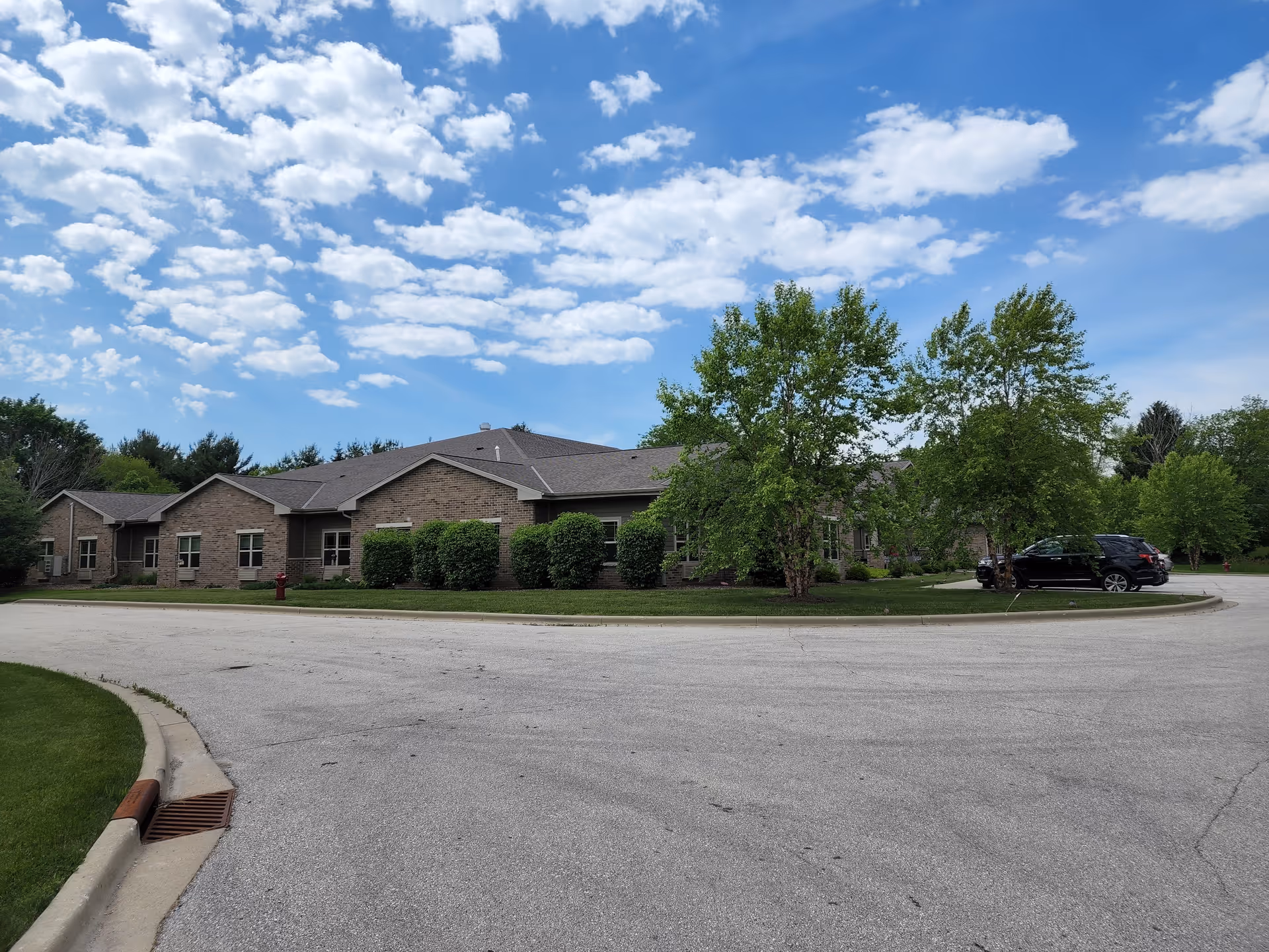 Exterior view of a single-story brick building with multiple windows, surrounded by green bushes and trees under a partly cloudy blue sky. There is a curved driveway or parking area in front with a black SUV parked on the right side.