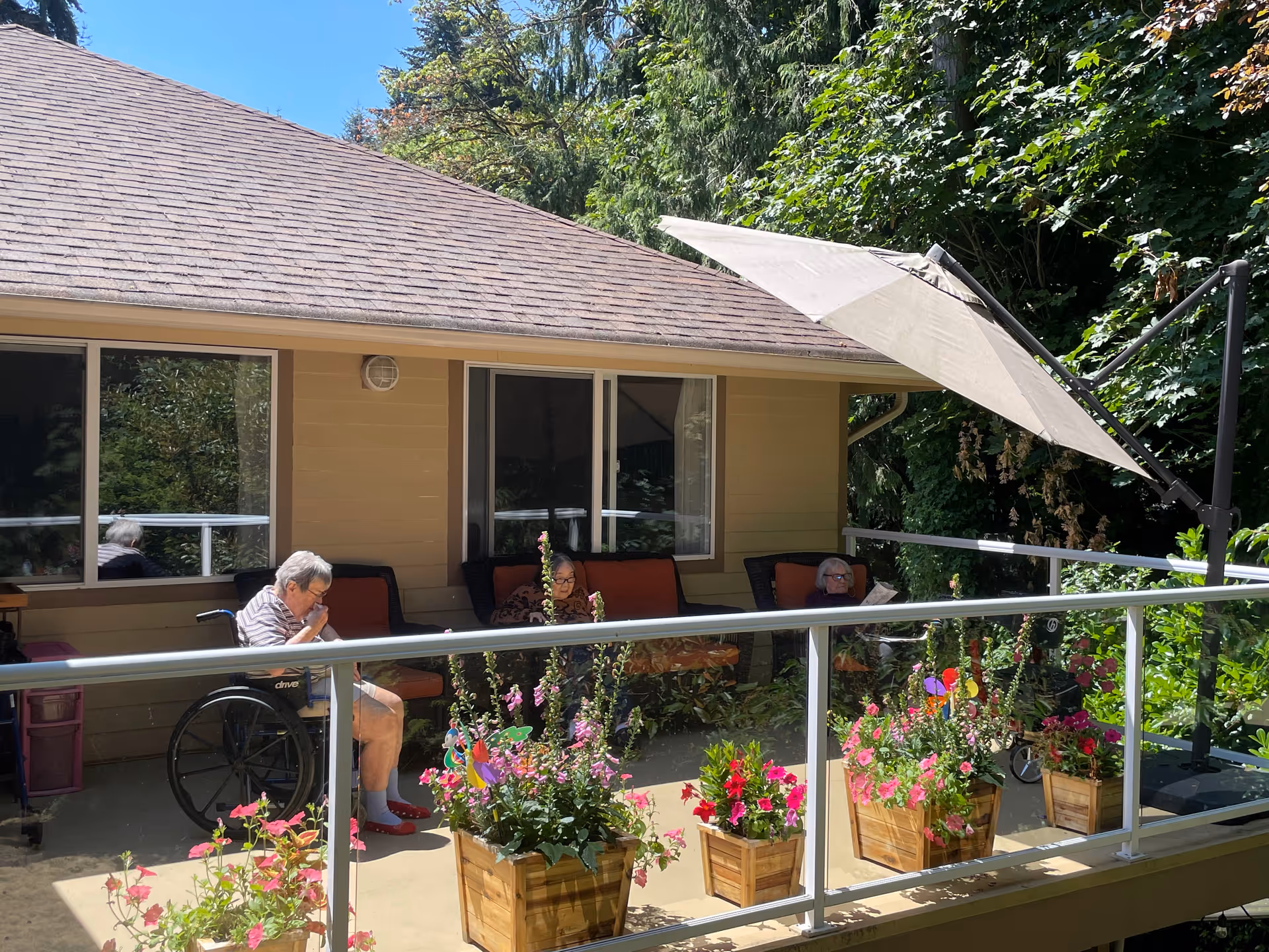Three people sit on a sunny outdoor patio with potted flowers, a glass railing, and a large umbrella in front of a single-story building.