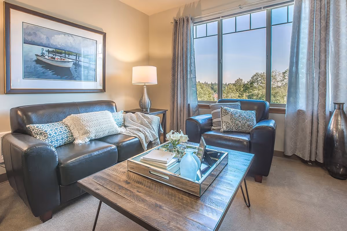A cozy living room with a dark leather sofa and armchair, decorated with patterned cushions and a throw blanket. A wooden coffee table with a mirrored tray holding books, a vase with flowers, and decorative items sits in front of the seating. A large window with curtains offers a view of trees outside, and a framed painting of a boat on water hangs on the wall. A table lamp on a side table provides warm lighting.