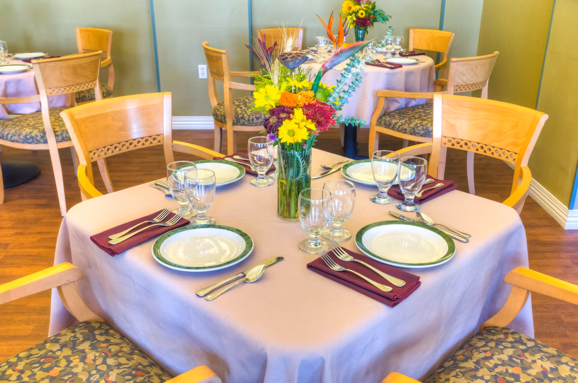 A dining room table set for four with white plates, silverware, glasses, and burgundy napkins on a light pink tablecloth. A vase with colorful flowers is in the center of the table. Wooden chairs with patterned cushions surround the table, and other similarly set tables are visible in the background.