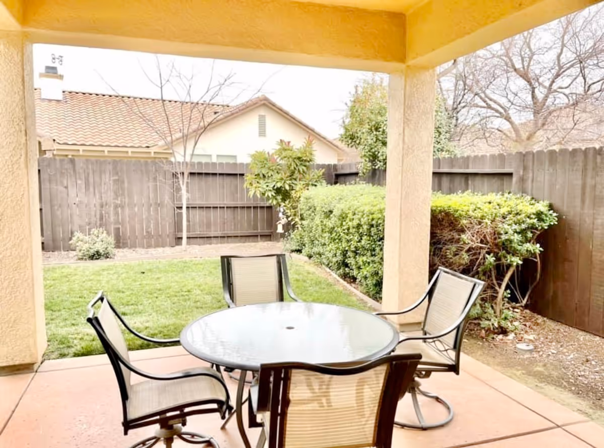 Covered backyard patio with a round glass table and four chairs overlooking a small lawn and fenced yard.