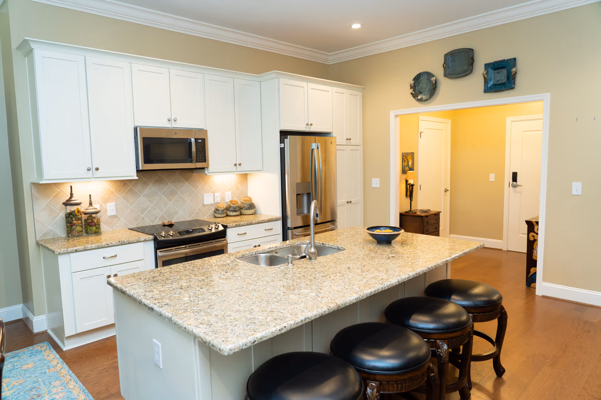 Modern kitchen with white cabinets, stainless steel appliances including a microwave and refrigerator, a large granite island with a sink and four black cushioned stools, and decorative plates on the wall above the doorway leading to a hallway.