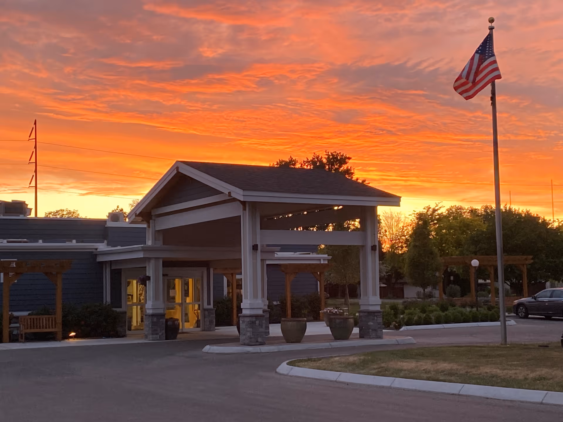 Entrance of Heatherwood Senior Living facility at sunset with a vibrant orange and pink sky, a covered drop-off area, an American flag on a flagpole, and surrounding greenery.