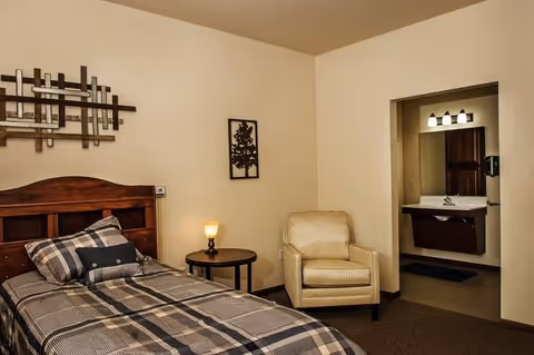 A furnished bedroom with a single bed dressed in plaid bedding, a nightstand with a lamp, an armchair, wall art, and a doorway leading to a bathroom sink.