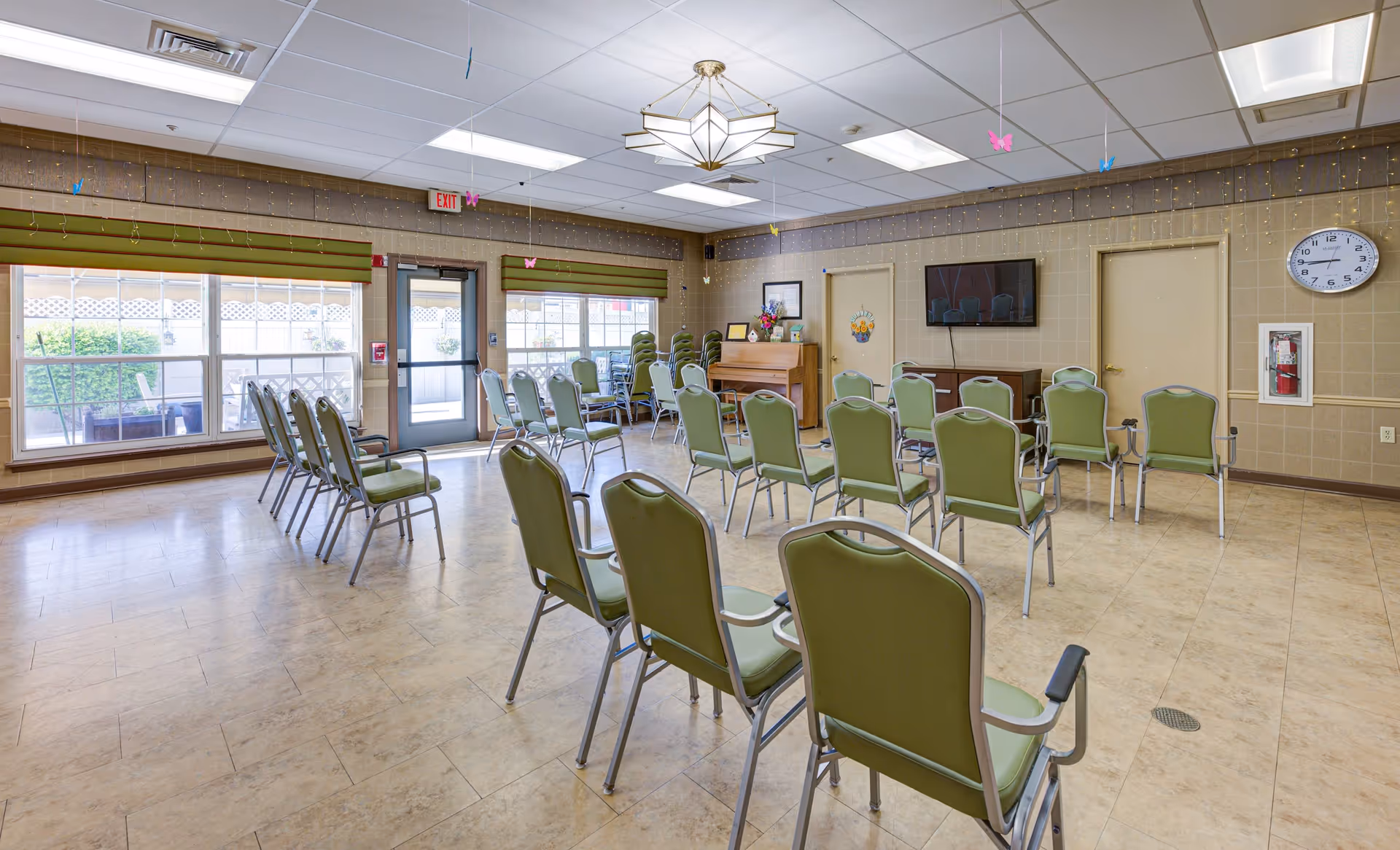 A spacious room with rows of green cushioned chairs arranged facing a wall-mounted flat screen TV. The room has large windows with green valances, a piano in the corner, a clock on the wall, and a door leading outside. The ceiling has white tiles with fluorescent lighting and decorative hanging butterflies.