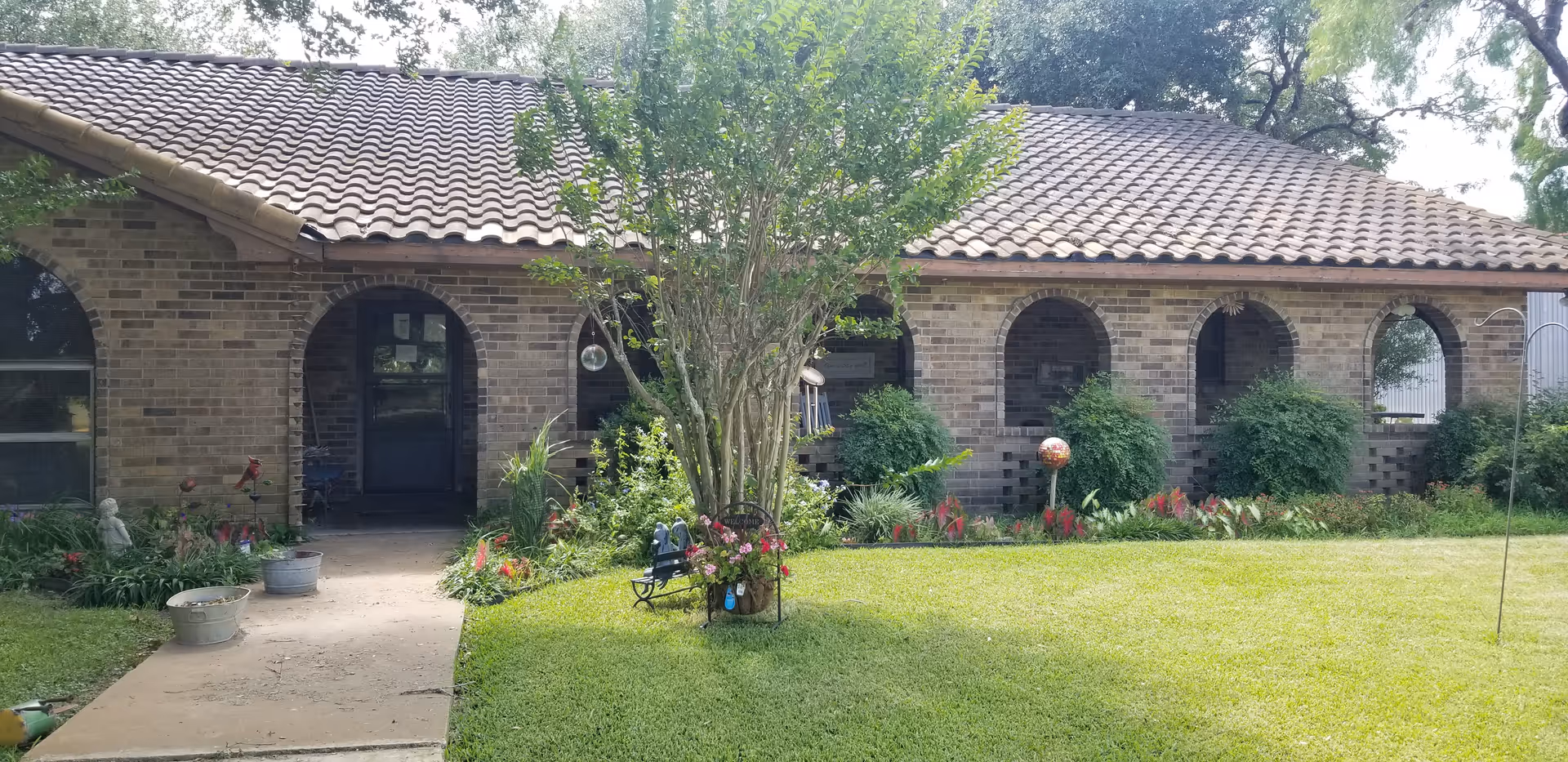 Front exterior view of a single-story brick building with a tiled roof, arched doorways and windows, surrounded by a well-maintained garden with green grass, bushes, flowers, and a small tree in the center.