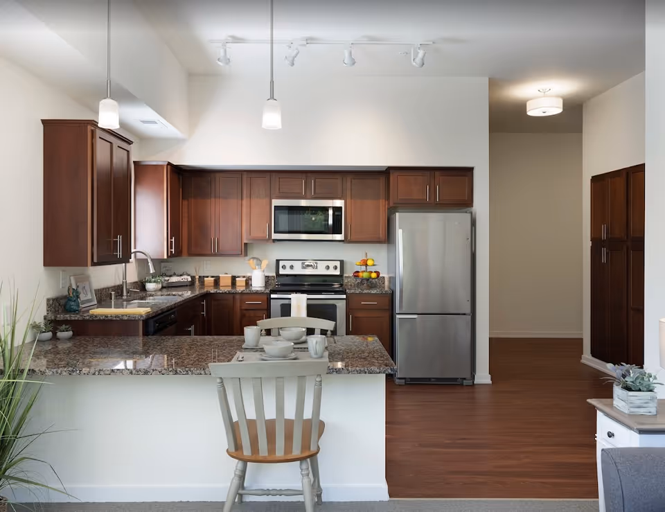 Modern kitchen with dark wooden cabinets, granite countertops, stainless steel refrigerator, stove, and microwave. A kitchen island with a chair and tableware is in the foreground. The floor is wooden, and there is a hallway visible to the right.