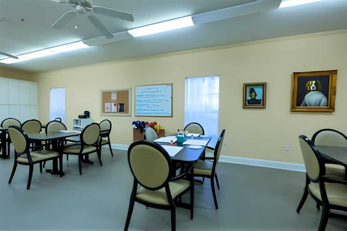 Bright communal dining/activity room with several tables and chairs, artwork on the walls, and overhead fluorescent lights.