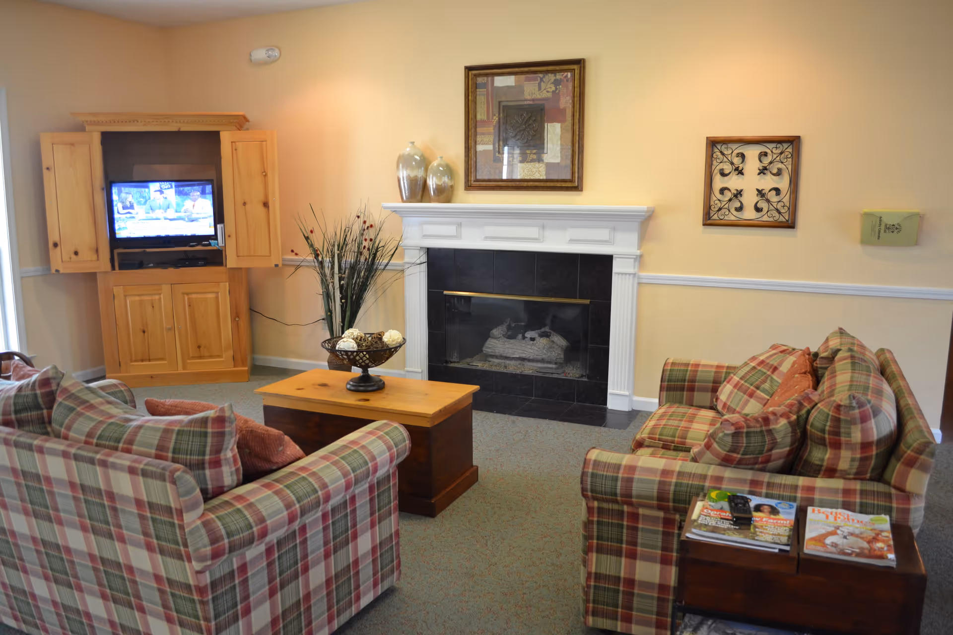 A cozy living room with two plaid sofas facing each other, a wooden coffee table in the center, a fireplace with a white mantel, decorative vases and framed artwork above it, and a wooden cabinet with a TV inside on the left side. There is also a small side table with magazines and a remote control.