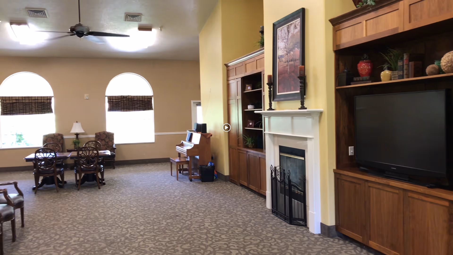 A spacious living room area with patterned carpet, two large arched windows with blinds, a table with four chairs, two armchairs, a piano with a bench, a white fireplace with a black screen, and wooden built-in shelves and cabinets. The shelves hold decorative items and a large flat-screen TV. The walls are painted yellow and beige.
