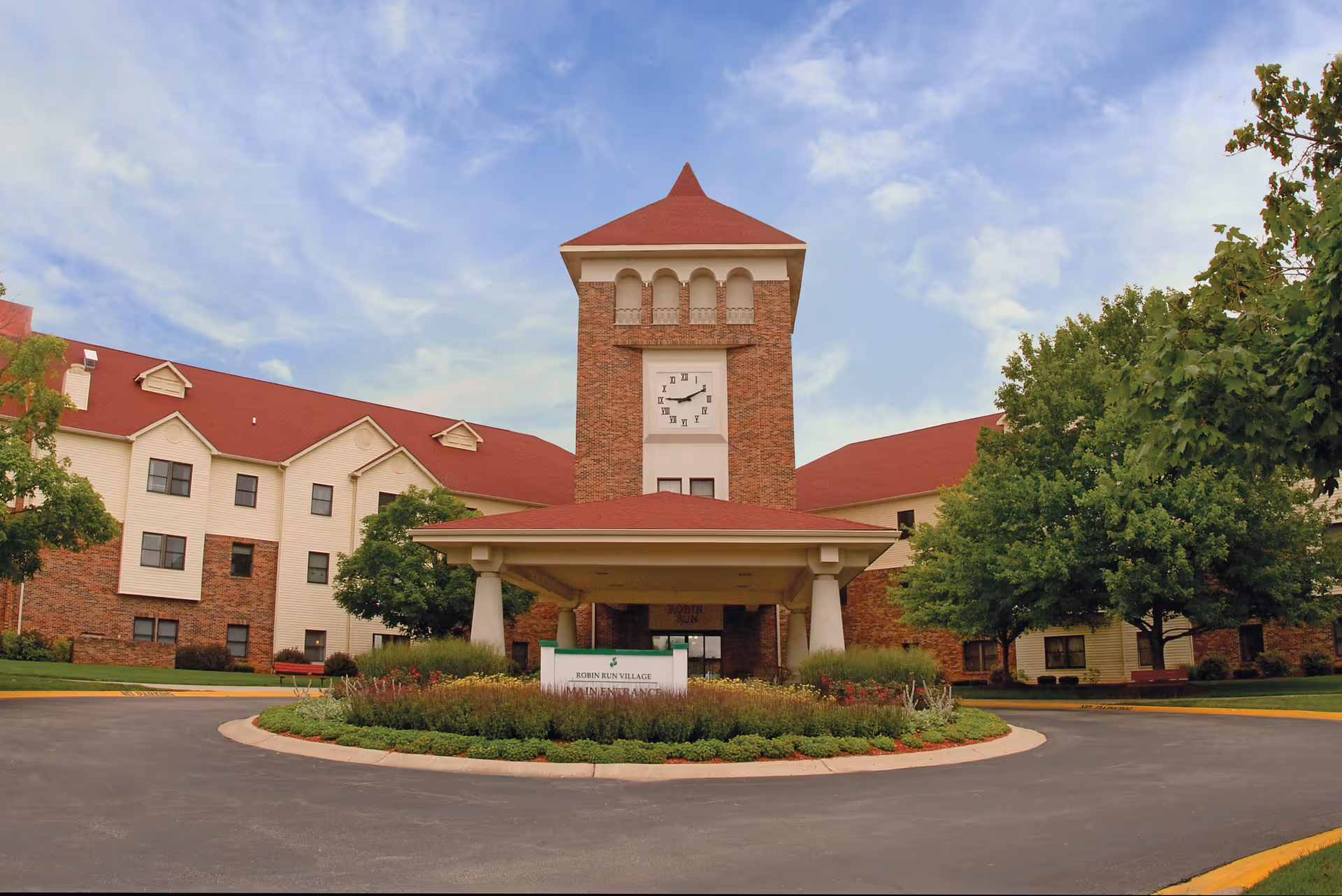 Front exterior view of Robin Run Village Senior Living facility featuring a large brick and beige building with a prominent clock tower and red roof, surrounded by green trees and landscaping under a partly cloudy sky.