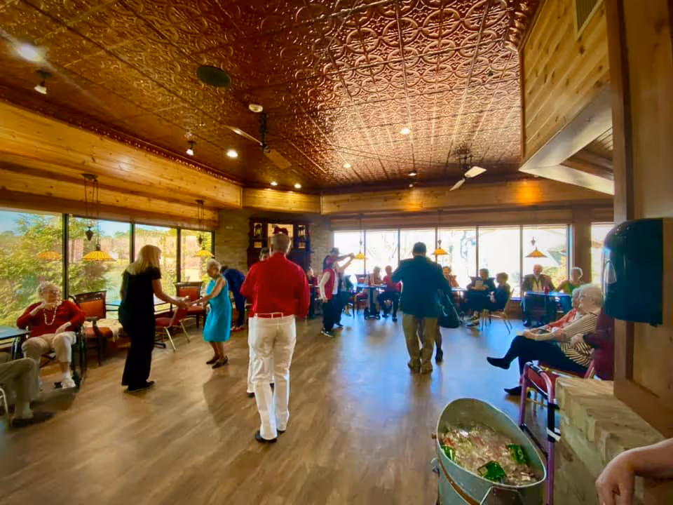 A spacious room with wooden walls and a decorative ceiling, filled with elderly people sitting and standing. Some are dancing or interacting in the center of the room, while others are seated along the walls near large windows letting in natural light. The atmosphere appears lively and social.