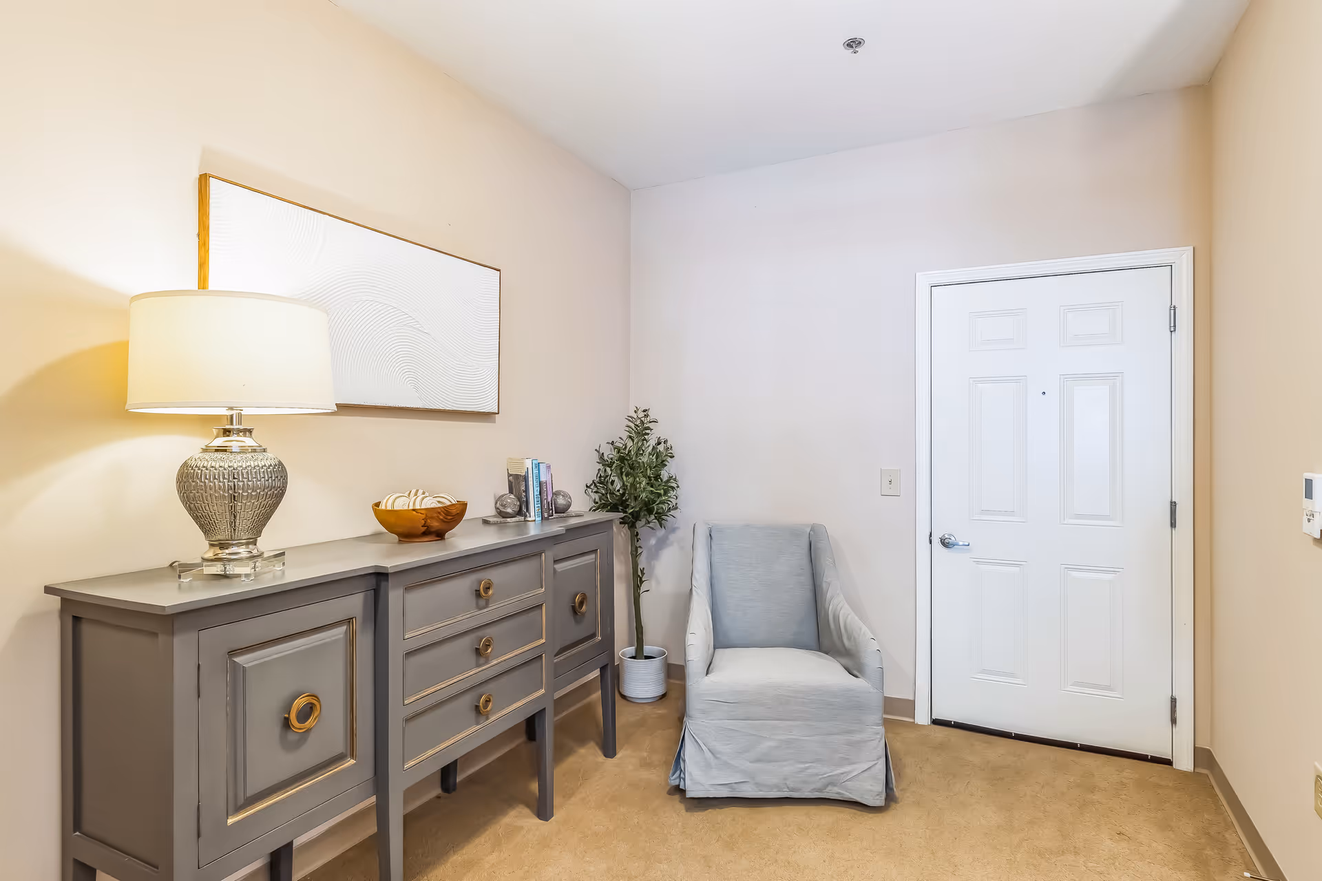 A small entryway area with a white door, a gray armchair, a gray sideboard with drawers and cabinets, a decorative lamp, a wooden bowl, some books, and a potted plant. The walls are light-colored and the floor is carpeted.