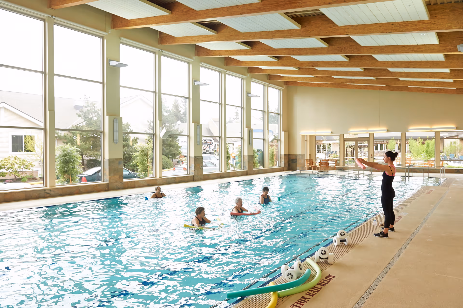 Indoor swimming pool with large windows letting in natural light. Four elderly women are in the pool participating in a water exercise class led by an instructor standing poolside. Pool equipment such as foam noodles and numbered floats are visible on the pool deck.