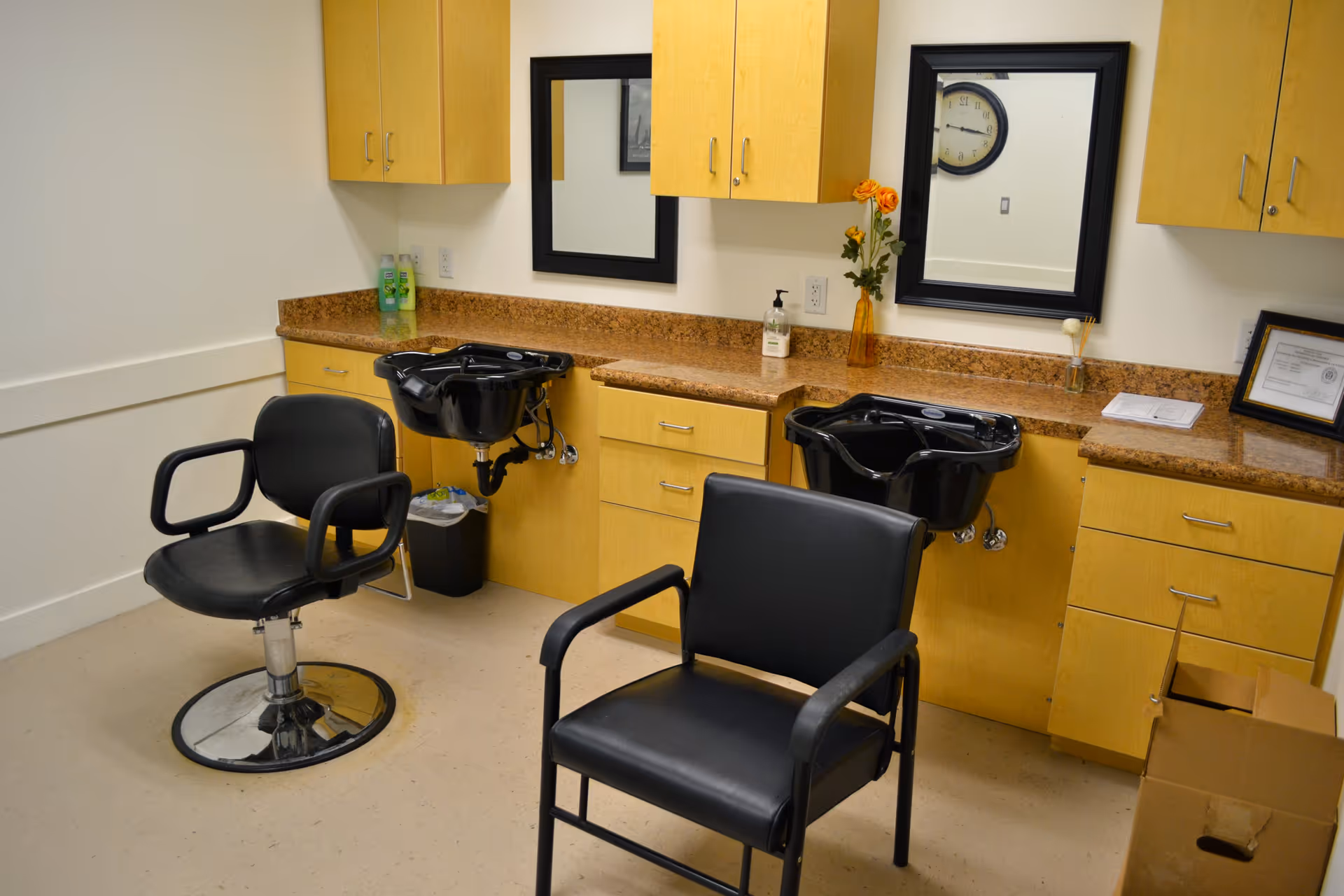 Interior view of a hair salon area in an assisted living facility with two black salon chairs, two black hair washing sinks, wooden cabinets, mirrors, and a countertop with a vase of flowers and hand sanitizer.
