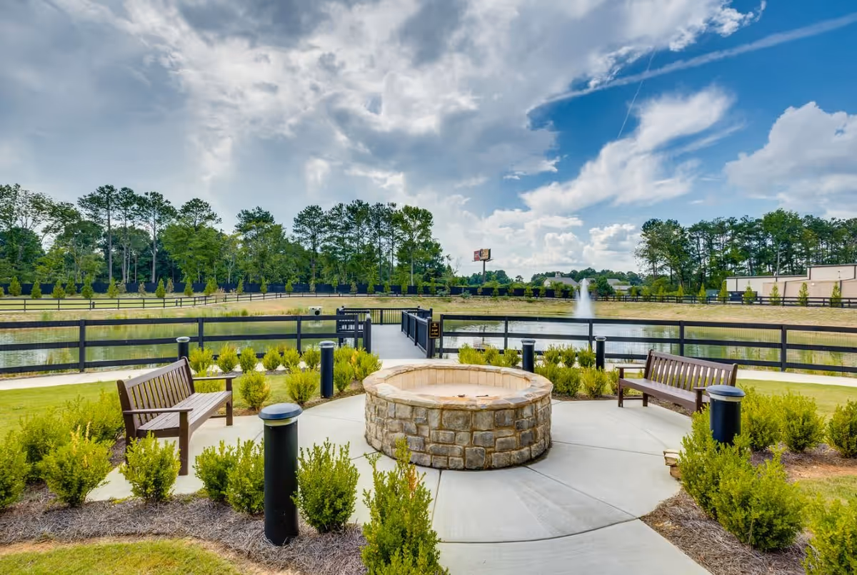Outdoor seating area with two wooden benches facing a circular stone fire pit, surrounded by small green bushes. In the background, there is a fenced pond with a water fountain and trees under a partly cloudy sky.