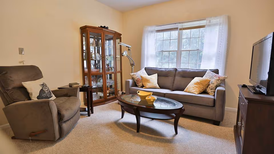 Bright living room with a gray sofa, recliner, glass coffee table, china cabinet and TV by a large window.