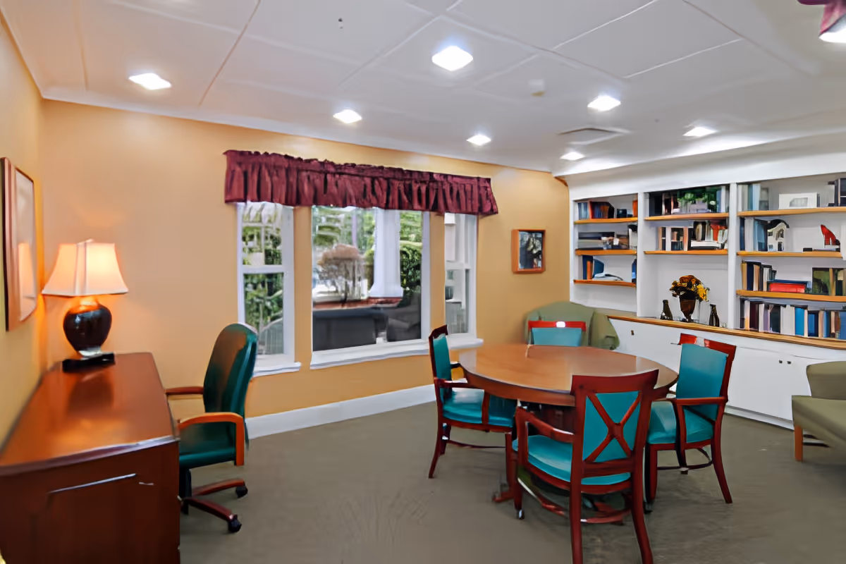 Bright common room with a round wooden table surrounded by teal-upholstered chairs, bookshelves along the back wall, and a window with a maroon valance.