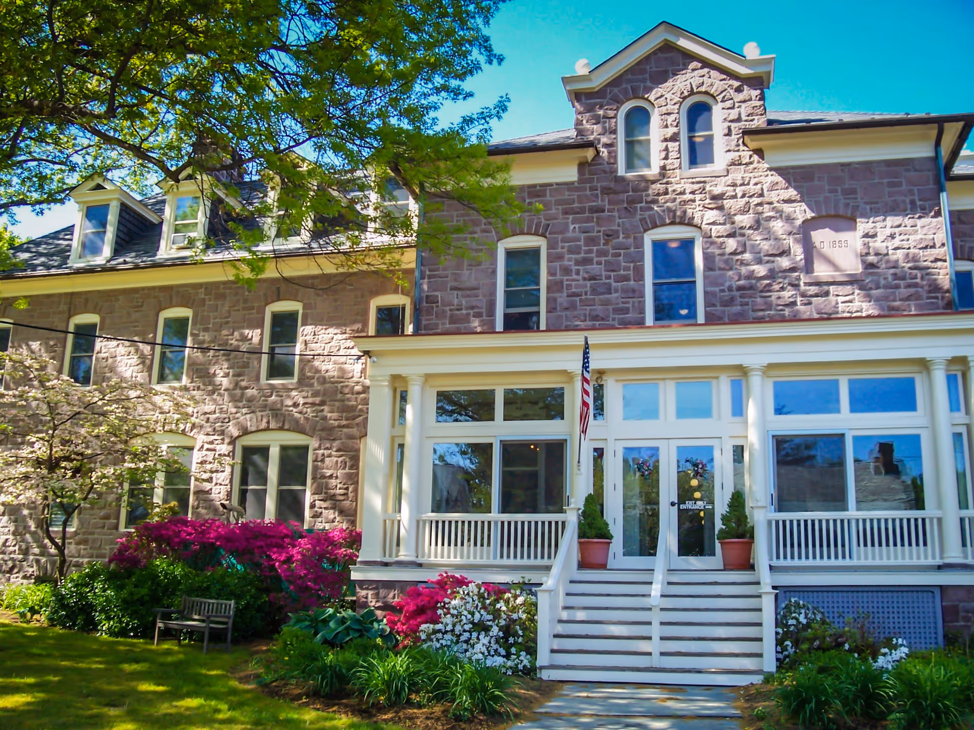 Front exterior view of a large stone building with multiple windows and a white porch entrance. There are stairs leading up to the porch with potted plants on either side of the door. The building is surrounded by green grass, colorful flowering bushes, and trees under a clear blue sky.