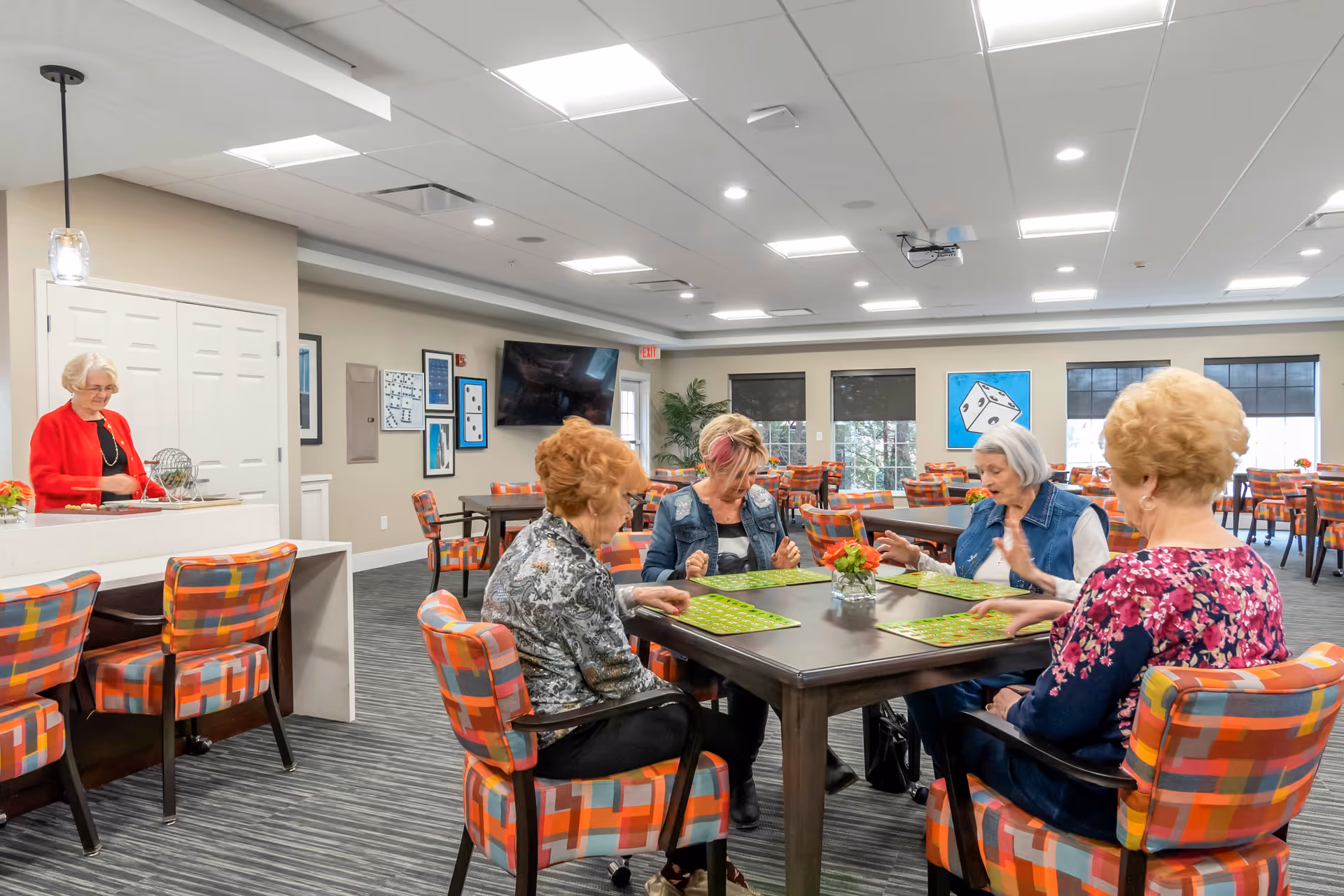 A group of elderly women sitting around a table playing a game with green cards in a brightly lit community room. Another elderly woman stands nearby at a counter. The room has colorful patterned chairs, large windows, and framed artwork on the walls.