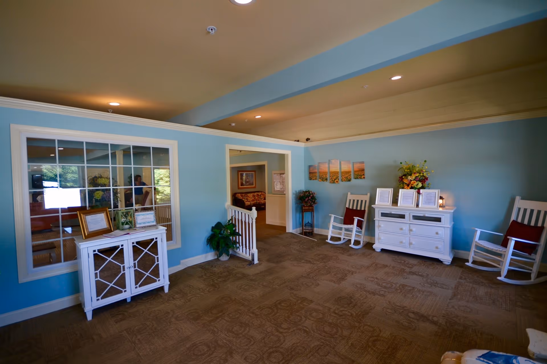 A cozy interior room with light blue walls and beige carpet. The room features two white rocking chairs with red cushions, a white dresser adorned with framed documents and a flower arrangement, and a small white cabinet with framed pictures on top. There is a large window with multiple panes looking into another room, and a doorway leading to a sitting area with a floral couch. The ceiling has recessed lighting and a beige color.