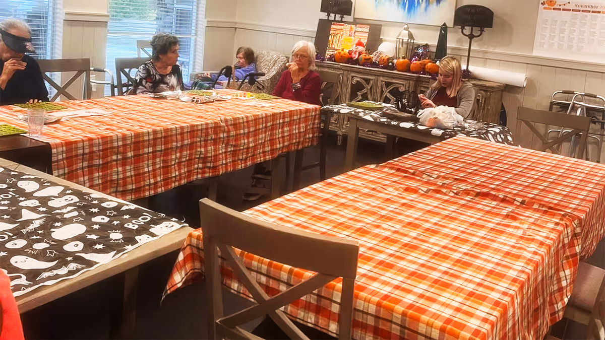 A group of elderly people sitting around tables covered with orange plaid and Halloween-themed tablecloths in a dining area. The room is decorated with small pumpkins and Halloween decorations on a sideboard against the wall. A calendar is visible on the wall, and a young woman is seated at one of the tables.