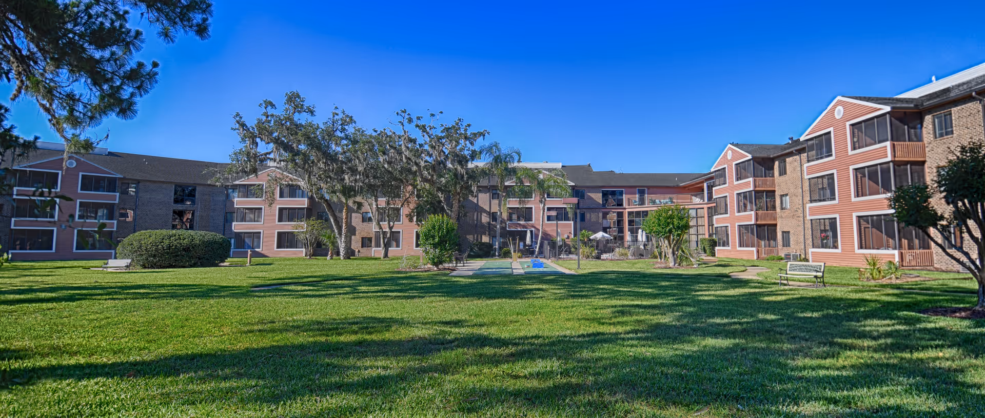 Well-maintained grassy courtyard with benches in front of a three-story brick senior living building under a clear blue sky.