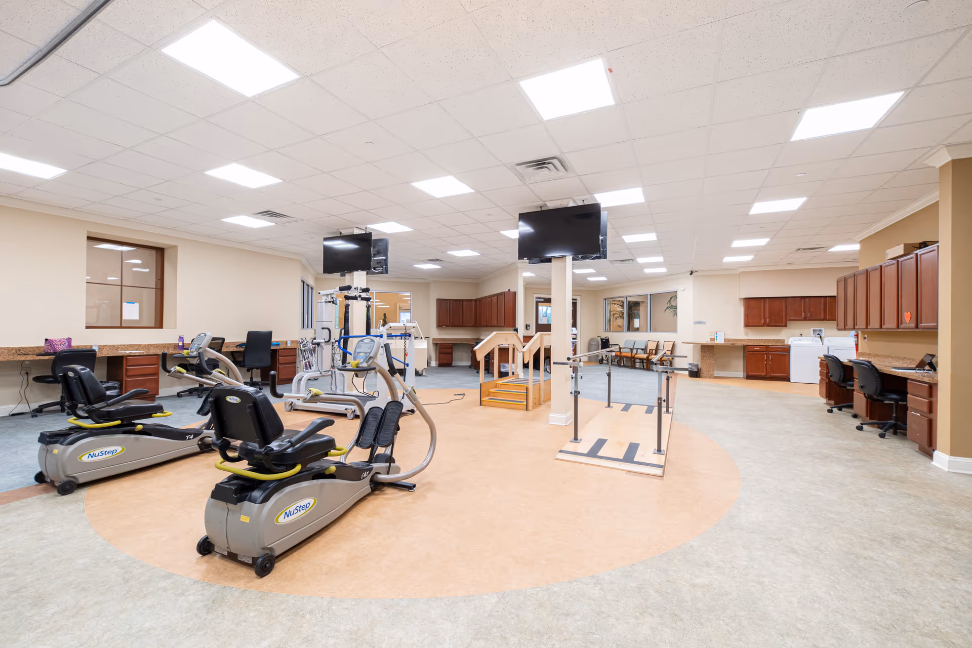A spacious rehabilitation or physical therapy room with exercise equipment including NuStep machines, parallel bars for walking practice, a small set of stairs with handrails, multiple desks with chairs, and two large wall-mounted TVs. The room has bright overhead lighting, beige walls, and a combination of carpet and vinyl flooring.