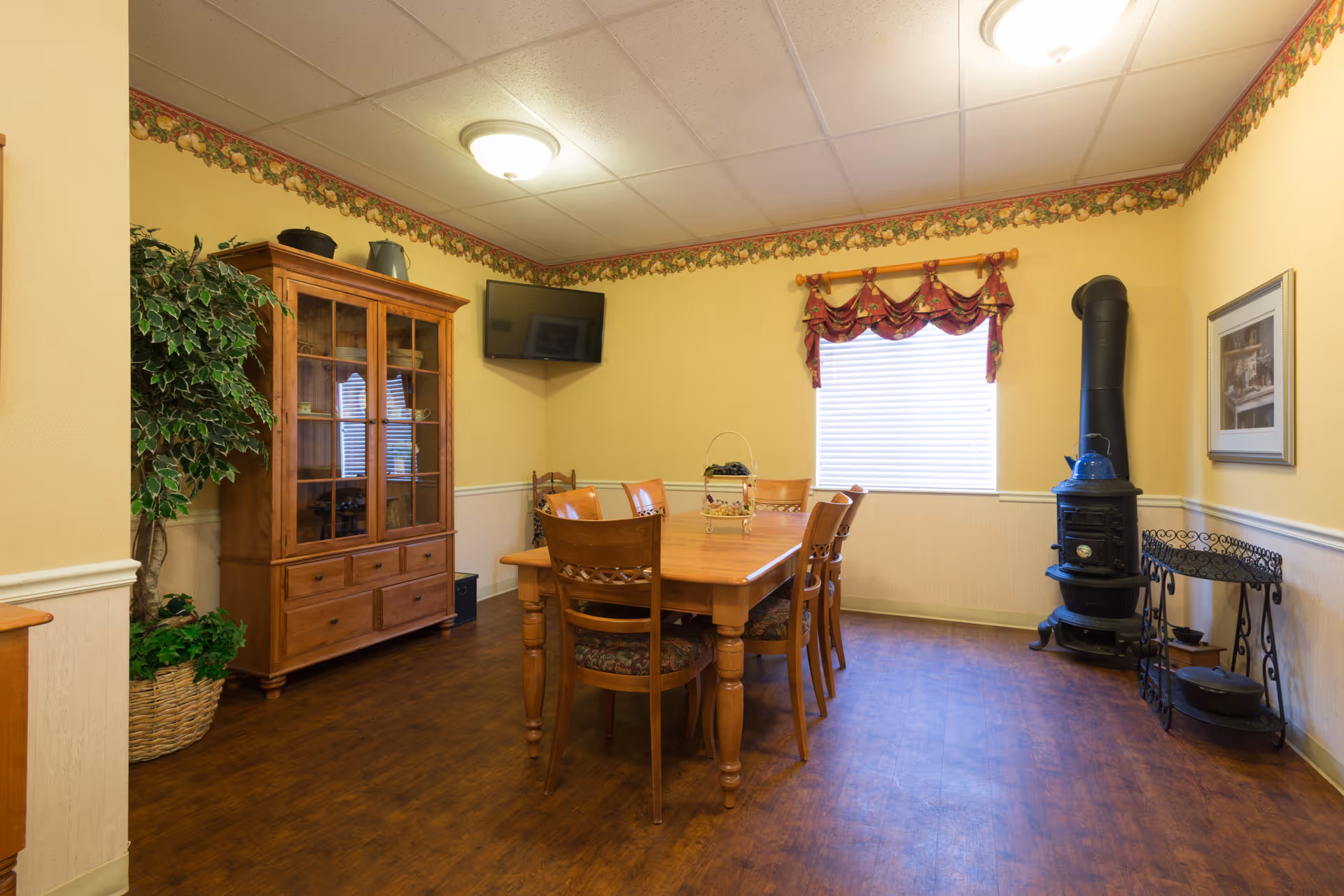 A cozy dining room with a wooden dining table and six chairs. The room has yellow walls with floral wallpaper border near the ceiling, a window with a red valance, a black wood-burning stove with a blue kettle on top, a wooden cabinet with glass doors, a potted plant, a wall-mounted TV, and a framed picture on the wall.