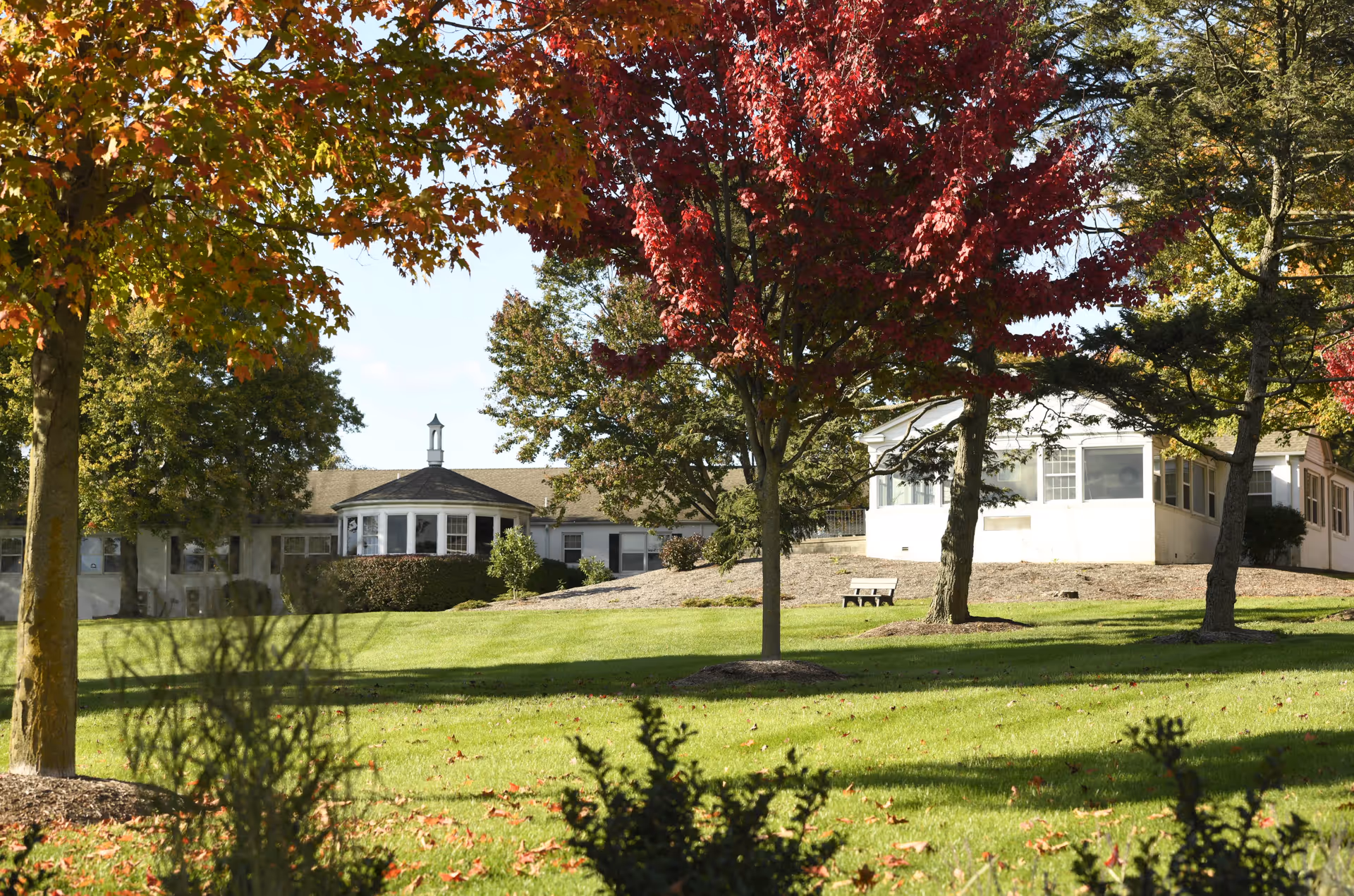 A grassy lawn with autumn-colored trees frames the front of a single-story nursing center building with a small gazebo-like bay and a bench.