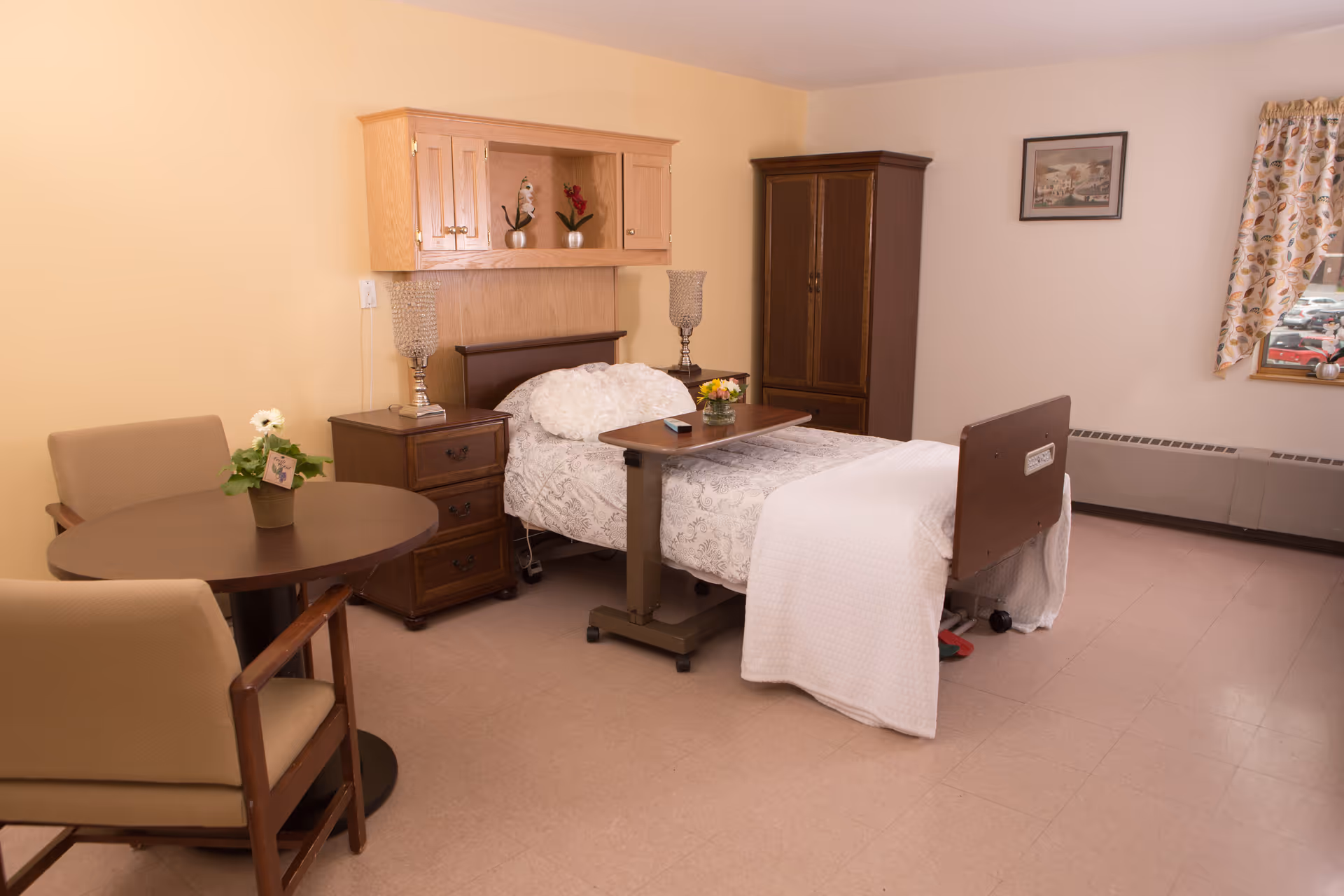 A neatly arranged bedroom in a senior living facility featuring a single hospital-style bed with white bedding and a wooden headboard. The bed has an overbed table with a small flower arrangement and a remote control on it. Next to the bed is a wooden nightstand with a lamp and a wooden cabinet. There is a small round table with two beige chairs and a potted plant on the table. The room has light-colored walls, a window with floral curtains, and a framed picture on the wall.