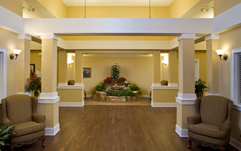 A spacious and well-lit interior common area with wooden flooring, beige walls, and white trim. The room features two striped armchairs on either side, several columns, wall-mounted light fixtures, and a central indoor garden with green plants and red flowers.