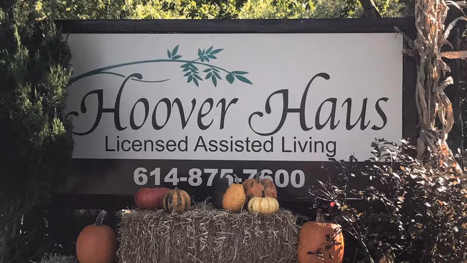 Outdoor sign reading "Hoover Haus Licensed Assisted Living" mounted above hay bales and pumpkins.