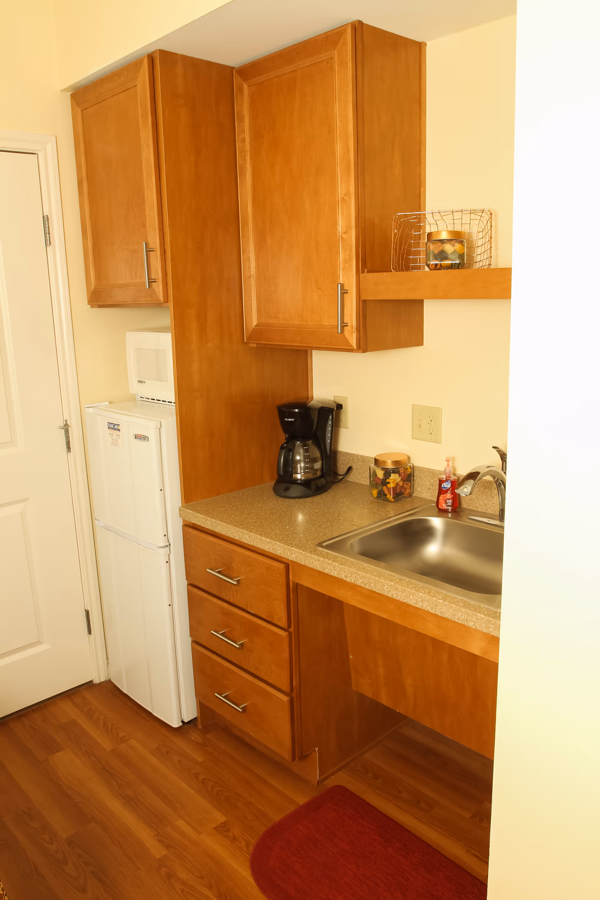 Small kitchenette with wooden cabinets, a countertop sink, coffee maker, mini fridge and microwave.